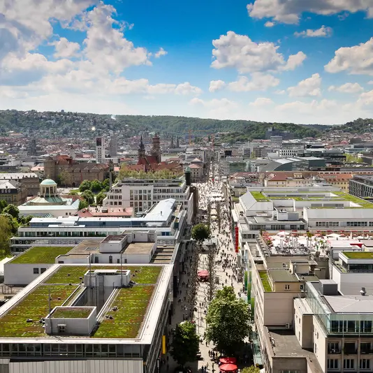 Luftaufnahme der Stuttgarter Innenstadt mit der belebten Königstraße, modernen Gebäuden und begrünten Dächern unter blauem Himmel.